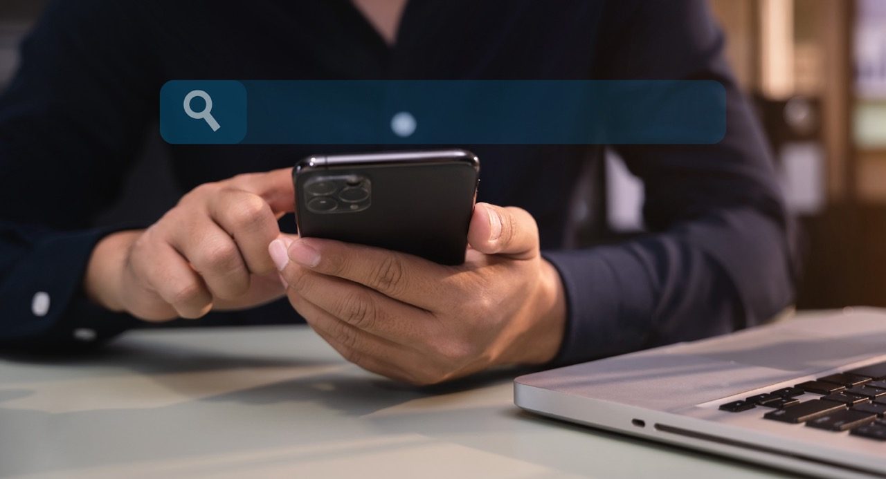 Homem pesquisando no celular apoiado em uma mesa de trabalho