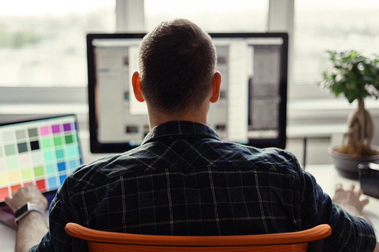 Designer trabalhando em dois monitores com uma cartela de cores ao lado